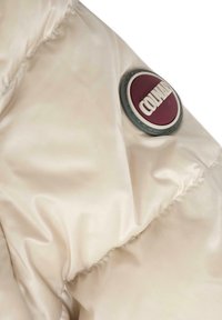 Dettaglio di un piumino beige con finitura lucida, caratterizzato da una rotonda patch logo bordeaux e bianco sulla manica. Texture liscia.