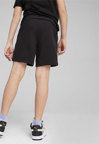 Shorts sportivi neri con tessuto liscio, vita elastica e dettagli di cucitura, abbinati a sneakers bianche e calzini azzurri chiari.