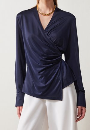 Femme portant un chemisier portefeuille manches longues bleu marine avec un devant drapé, associé à un pantalon blanc et un collier épais en or.