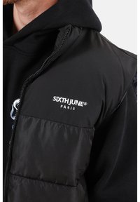 Zwarte gewatteerde vest met een ritssluiting aan de voorkant en het logo "SIXTH JUNE PARIS" in witte borduursel. De binnenvoering lijkt zacht en vormt een contrast met de buitenste textuur.