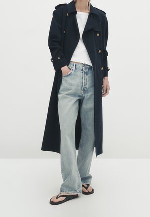 Personne portant un long manteau noir avec des boutons dorés, un haut blanc, un jean large bleu clair et des sandales noires, debout avec une main dans la poche.