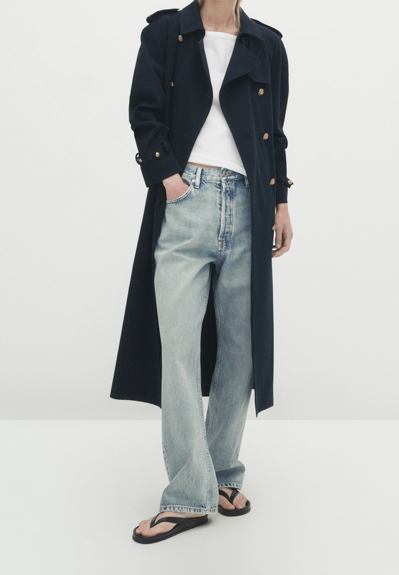 Personne portant un long manteau noir avec des boutons dorés, un haut blanc, un jean large bleu clair et des sandales noires, debout avec une main dans la poche.