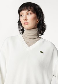 Witte oversized trui met een V-hals, gestructureerd breiwerk, met een klein groen krokodillenembleem op de linkerborst. Gelaagd over een beige coltrui.