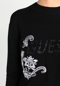Svart stickad tröja med metallisk "GUESS"-tryck och vit broderad blommönsterdesign med intrikata detaljer på framsidan.