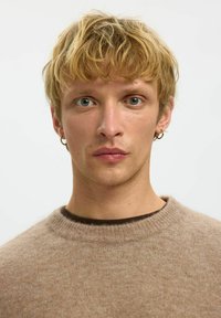 Beige gestrickter Pullover mit runder Halslinie, weicher Textur, über einem dunkelbraunen Unterhemd getragen; das Modell hat kurzes, zerzaustes blondes Haar.