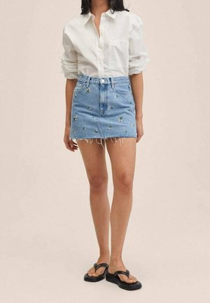 Jupe en denim bleu clair avec broderies florales, ourlet effiloché, taille haute et poches avant, associée à une chemise blanche à boutons et des tongs noires.