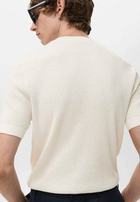 Suéter de punto acanalado en color crema, de manga corta y ajuste relajado, con un cuello redondo y una textura suave. Combinado con pantalones oscuros.