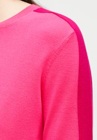 Κοντινό πλάνο ενός ροζ πλεκτού πουλόβερ που δείχνει τον ώμο και το πάνω μέρος του βραχίονα με λεπτομέρεια διχρωμίας σε πιο σκούρο ροζ.