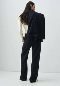 Costume deux pièces avec un tissu rayé sombre. Blazer ajusté drapé sur l'épaule, associé à un pantalon large. Haut en tricot léger en dessous.