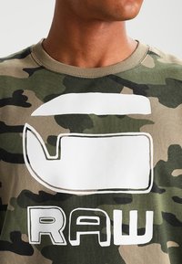 Maglione a motivo mimetico in tonalità di verde e marrone, con un grande dettaglio grafico bianco e la scritta "RAW" in bold bianco.