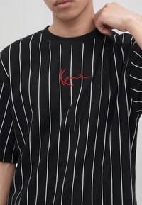 Persona con camiseta negra con rayas blancas verticales y texto rojo bordado en el pecho, ajustando el cuello con una mano.