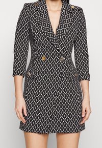 Frau in einem schwarzen Kleid mit weißem geometrischem Muster, goldenen Knöpfen, Dreiviertelärmeln und Reverskragen, die Hände entspannt an den Seiten.