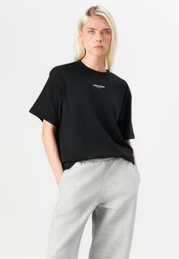 Black cotton t-shirt with short sleeves, relaxed fit, featuring a small white logo on the chest. Paired with light grey sweatpants.