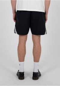 Homme portant un short noir avec des bandes latérales blanches et grises, des chaussettes blanches montantes et des chaussures en cuir noires, debout devant un fond clair uni.