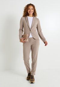 Costume ajusté marron clair avec une veste cintrée et un pantalon assorti, porté sur un t-shirt blanc, associé à des baskets décontractées marron.