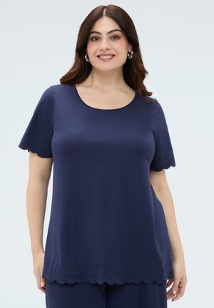 Donna sorridente con capelli scuri che indossa una maglietta blu navy a maniche corte e pantaloni con bordi smerlati e ricamo floreale sottile.