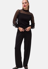 Musta haalari, jossa on vartalonmyötäinen yläosa, jota koristaa aaltoileva pitsikerros ja pitkät, läpinäkyvät hihat. Mallia täydentävät levenevät housut.