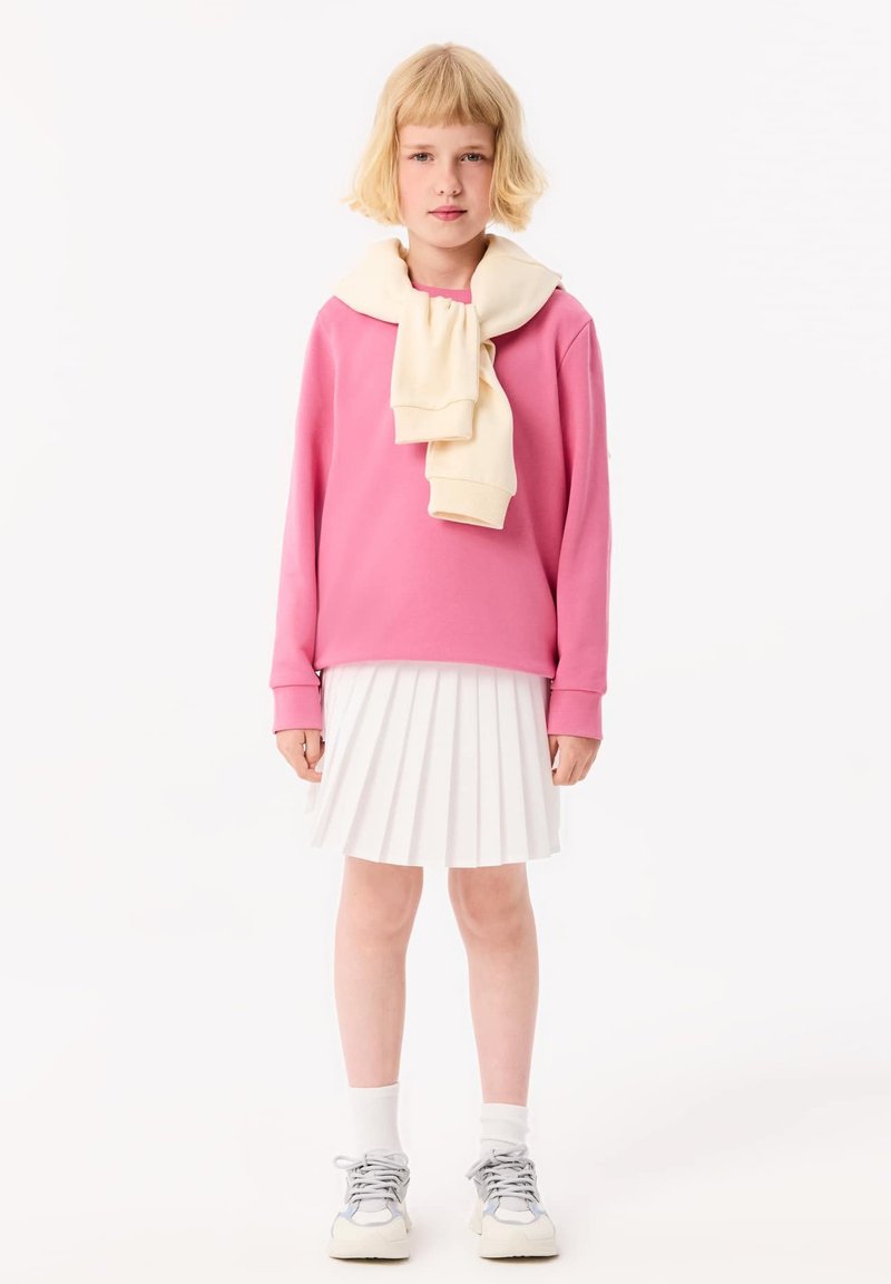 Rosa Sweatshirt mit einer cremefarbenen Kapuzenjacke um den Hals drapiert, kombiniert mit einem weißen, plissierten Rock und grauen Sneakern. Einfaches Design, lässiger Stil.