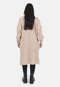 Cardigan lavorato a maglia beige con maniche lunghe, caratterizzato da una silhouette ampia, texture a coste e lunghezza fino a metà polpaccio, abbinato a leggings neri e stivali.
