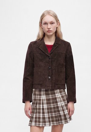 Jeune femme porte une veste boutonnée marron foncé par-dessus un haut rouge et une jupe plissée à carreaux marron et blanc sur un fond uni.