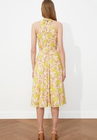 Robe florale avec un col halter, des couleurs pastel et une jupe évasée. Le tissu présente un mélange de fleurs vertes et oranges sur un fond clair.