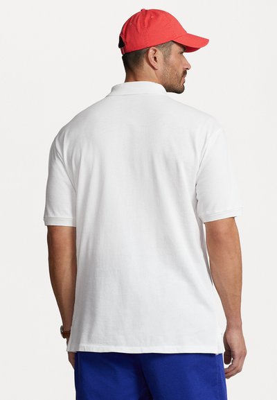 White polo shirt with short sleeves, featuring a straight hem and subtle side slits. Worn with a red cap and blue shorts.