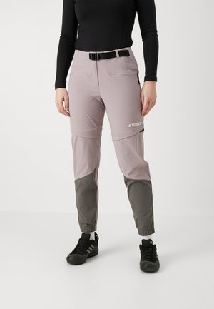 Osoba nosząca jasnofioletowe i szare spodnie outdoor TERREX, czarną koszulkę z długim rękawem oraz czarne sneakersy, stojąca na białym tle.