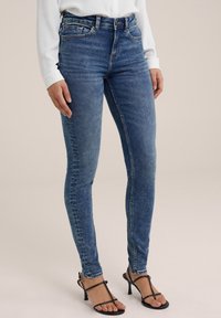 Jeans ajustados de mezclilla azul de cintura alta con un diseño clásico de cinco bolsillos, un ligero desgaste y una textura suave; combinados con tacones negros de tiras.