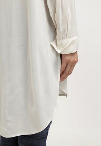 Main portant une chemise blanche à manches longues avec poignet boutonné, détail de fente latérale, sur un jean foncé sur fond uni.