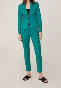 Turkusowy dopasowany garnitur z dopasowaną marynarką na jeden guzik, pasującymi zwężanymi spodniami i czarnymi klapkami. Gładka faktura materiału.