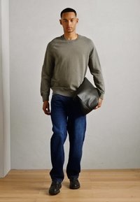 Sweatshirt gris avec un col ras du cou, jeans bleus et chaussures noires. Homme tenant un sac texturé gris foncé, debout sur un sol en bois clair.