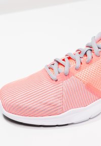 Chaussure de sport avec une tige en corail texturée et côtelée, dotée de lacets et d'accents gris. Semelle blanche incurvée avec un motif de traction noir.