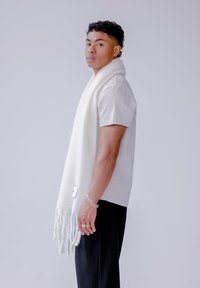 Witte sjaal met franjes hangt over een grijs T-shirt met korte mouwen, gecombineerd met zwarte geplooide broek. De sjaal heeft een klein label onderaan.