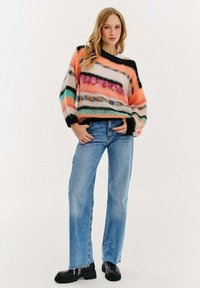 Wielokolorowy sweter dziergany z poziomymi paskami w kolorach różowym, pomarańczowym, czarnym i turkusowym. Połączony z jasnoniebieskimi spodniami typu straight-leg i czarnymi butami.