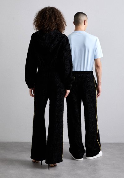 Ensemble de survêtement noir texturé à capuche avec impression de logo visible, associé à un t-shirt bleu clair et un pantalon noir avec des rayures jaunes sur les côtés.