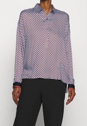 Camisa de botones de manga larga en tejido azul claro con un patrón circular rojo. Puños en contraste en azul marino y un ligero detalle de dobladillo asimétrico.