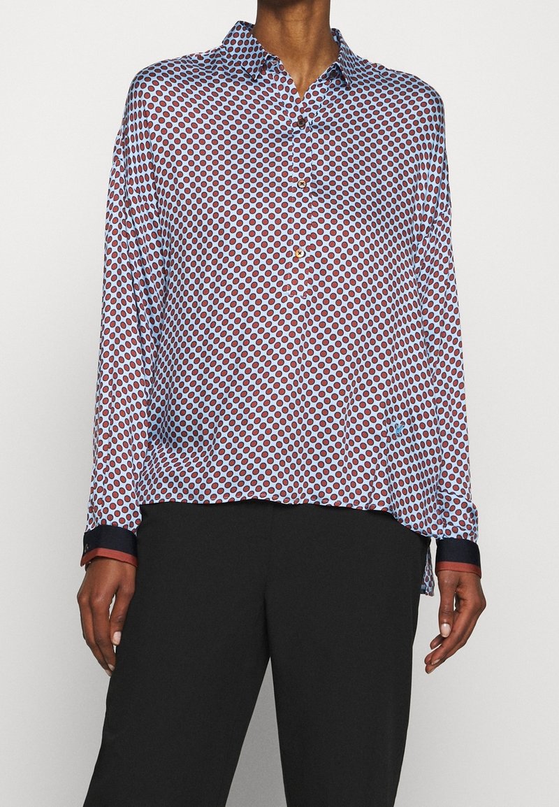 Long-sleeve button-up shirt in light blue fabric featuring a red circular pattern. Contrast navy cuffs and slight high-low hem detail.