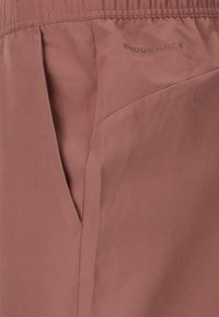 Staubige rosa, leichte Shorts mit einem elastischen Bund, einer Seitentasche und einem Logo, das in einem dunkleren Farbton gedruckt ist. Glatte Textur und minimalistisches Design.
