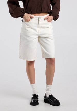 Person trägt weiße kniehohe Shorts, ein kurzes braunes Langarmshirt, weiße Socken und schwarze Loafers und steht mit den Händen in den Taschen.