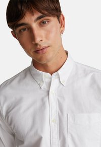 Camicia bianca con bottoni, caratterizzata da un colletto classico e una tasca sul petto, realizzata in morbido cotone con una texture delicata.