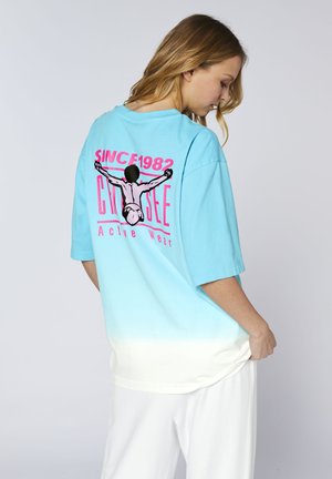 Junge Frau trägt ein blau-weißes Verlaufs-T-Shirt mit schwarzer Grafik und pinker Schrift "Since 1982 Cruise Active Wear" auf dem Rücken.