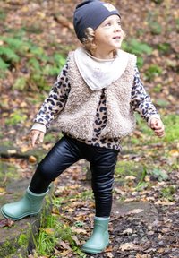 Enfant portant une chemise à manches longues à imprimé léopard, un gilet en fausse fourrure beige, des leggings noirs et des bottes en caoutchouc vertes dans un cadre forestier.