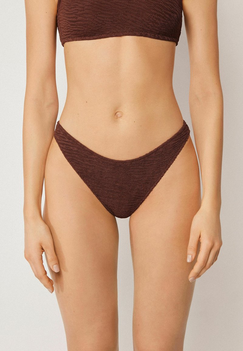 Buste féminin portant un haut de bikini texturé marron foncé et un bas de bikini assorti à taille haute, sur un fond clair uni.