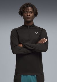 Νέος άνδρας με σταυρωμένα χέρια, φοράει μαύρο μακρυμάνικο μπλουζάκι με φερμουάρ Puma και τιρκουάζ παντελόνι, με φόντο γκρι.