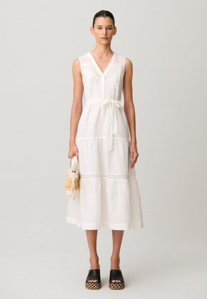 Donna che indossa un vestito midi bianco senza maniche con cintura annodata in vita, tiene una piccola borsa intrecciata e porta sandali platform neri con suola intrecciata.