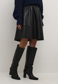 Jupe A-line en cuir noir avec taille élastique, associée à des bottes noires à talons hauteur genou. Pull en tricot bleu texturé.