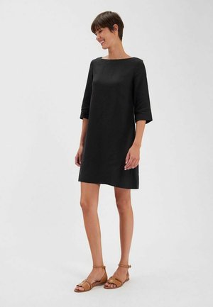Robe en lin noir avec un col bateau et des manches trois-quarts, coupe ample, longueur mi-cuisse, associée à des sandales plates beiges.