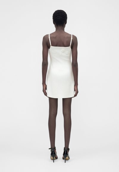 Γυναίκα που στέκεται με την πλάτη προς τα πίσω, φοράει λευκό μίνι αμάνικο φόρεμα με κεντήματα λουλουδιών και μαύρα ψηλοτάκουνα παπούτσια με απλό φόντο.