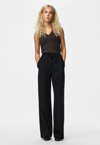Joven mujer con cabello rubio largo y rizado, vestida con una camiseta negra sin mangas y pantalones negros sueltos, de pie con las manos en los bolsillos sobre fondo blanco.