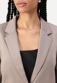 Beige blazer med skarpa slag, strukturerad tyg och en figursydd silhuett. Buren över en svart topp, som visar en minimalistisk design.